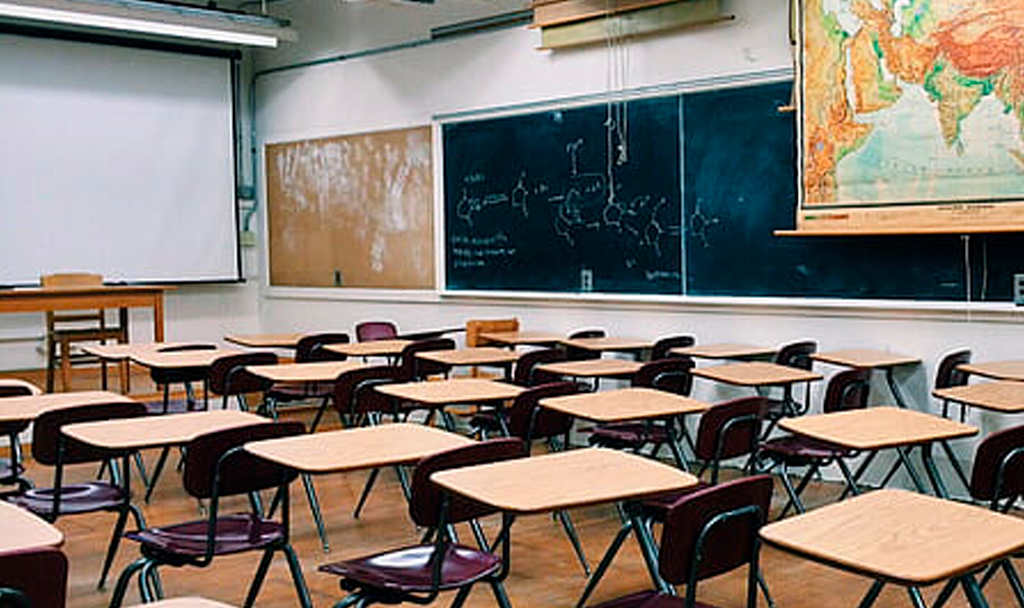 a traditional looking classroom with empty seats a map of the globe, a projector screen, and blackboards around the room.