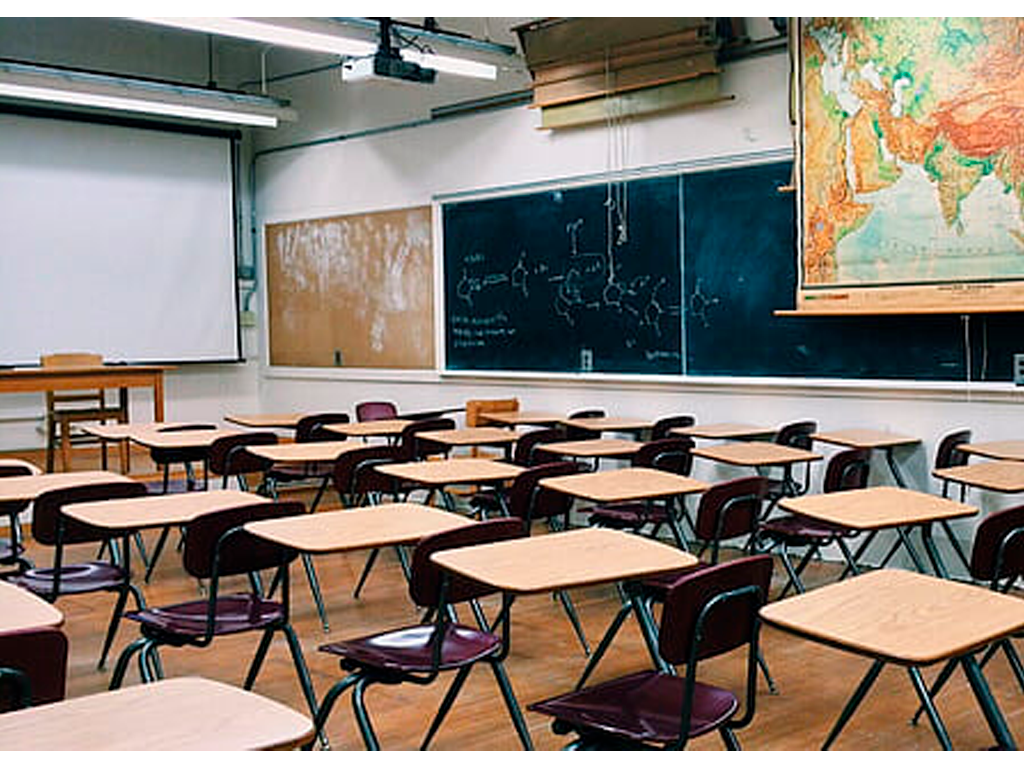 a traditional looking classroom with empty seats a map of the globe, a projector screen, and blackboards around the room.